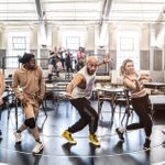 In a brightly lit room, five people are dancing in a line. A number of other people sit on a raised platform behindf them.