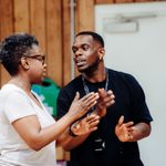 Two people having a conversation in a rehearsal room, gesturing with their hands. The person on the left is a woman wearing a white t-shirt and glasses. The person on the right is a man wearing a black t-shirt and an ID lanyard.