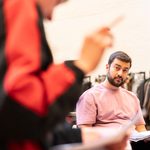 An actor in rehearsals, sat down and holding a script. He has short brown hair and is wearing a pink t-shirt. He is listening to someone in a red and black jacket whose arm is out of focus in the left of the image.
