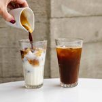 A person pouring coffee from a small cup into a tall glass filled with ice and milk, creating an iced latte. Next to it is a tall glass of iced black coffee, both placed on a white surface with a stone wall background.