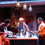 Three men gather around a kitchen island, focusing intently on cooking tasks. One man on the puts chicken wings on a grill, the man in the centre wears a yellow bandana, and the man on the right tends to a pot. Above them, three pendant lights illuminate the scene.