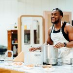 A man wearing a white apron and black tank top stands at a kitchen counter in a rehearsal room, smiling. There are various pots and pans on the counter as well as little dishes with chicken wings.