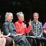 In front of a black background, a panel of five people sit in a row, the central figure holds a microphone addressing an audience who are out of shot to the right, the rest of the panel smile and laugh.