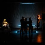 To the left, a man sat on a bed, holding a phone to his ear. A woman's face is projected onto his chest. To the right, a woman stands facing to the side, on the phone and gesturing with her left hand. In the middle, three people stand in shadow, looking at something on a table.
