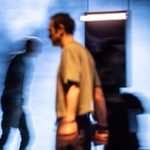 Blurred image of two people walking in a dimly lit, foggy environment. One person is more in focus, lit with orange light, while another is more shadowy and less distinguishable. A bathroom mirror with a rectangular light hangs in the background.