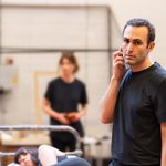 An actor in rehearsals, holding a phone to his right ear and looking off to the side with a serious expression. He has short black hair and is wearing a black t-shirt.
