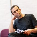 An actor in rehearsal, leaning on his elbow and resting a script on his knee. He has short black hair and is wearing a black t-shirt.