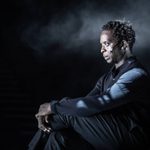 Kobna Holdbrook-Smith as Dr Toby Sealey in The Effect. He is wearing all black and sitting in a chair with his arms crossed.