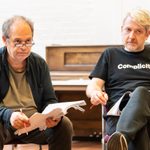 Two men in casual clothes sat in a rehearsal room, holding scripts and pencils. They are both looking ahead at something.
