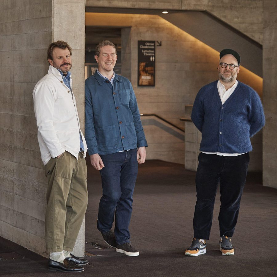 Lasdun-Jon-Rotheram-Tom-Harris-John-Ogier-Marksman-masthead Three men stand together and smile at the camera. One wears a white shirt, one a blue shirt and the other a navy cardigan. The backdrop is concrete walls and the top of a staircase.
