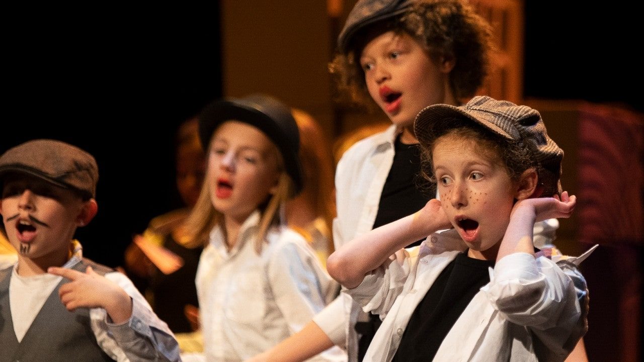 2019LetsPerform-flatcaps Four children onstage performing in a play, wearing flat caps and with painted freckles and a moustache.