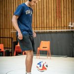 A young man wears over ear headphones, he is looking down at the ball he is playing keepy up with. He has afro hair which is shaved at the side but the top is long and peroxide dyed on the top. He wears a dark blue tshirt which says Utah State across the chest. He wears black jogger style shorts, he has white sport socks up to the ankle which have an orange band around them. He wears blue trainers.