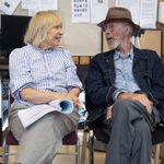 Two people are sitting next to each other on chairs in a casual setting. The woman, with blonde hair and wearing a striped shirt, holds papers and smiles, while the man, with a white beard, denim jacket, and hat, looks at her and talks.