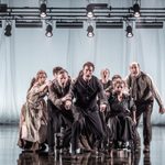 An ensemble of actors, clustered in a group with hands on each other's shoulders. Their expressions are tense and mouths open.