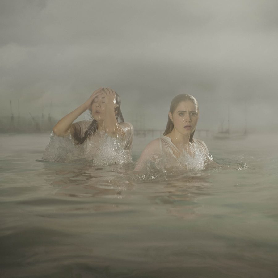 London-Tide-National-Theatre-2000x1000 Two women in thin white dresses stand in a river, soaking wet, one is pushing her hair back from her face. A low-lying city skyline and bridge are behind them in the distance.