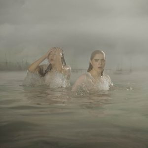 London-Tide-National-Theatre-2000x2000 Two women in thin white dresses stand in a river, soaking wet, one is pushing her hair back from her face. A low-lying city skyline and bridge are behind them in the distance.
