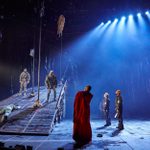 A dramatic theater scene with actors on stage. One actor in a red robe stands at the center while others in military attire are positioned on an inclined platform and on the ground. Blue lighting creates a tense atmosphere.