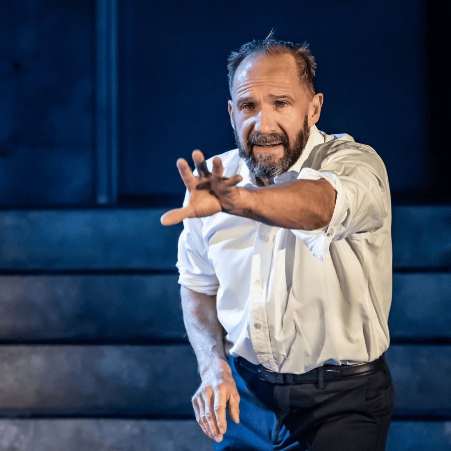 Macbeth-Ralph-Fiennes-National-Theatre-at-Home-masthead A man (Ralph Fiennes) with a beard and short hair, wearing a white shirt with rolled-up sleeves, stands on stage. He extends one arm forward, with a serious expression. The background is dimly lit with concrete steps visible.