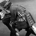 Two actors in period military costumes perform a dramatic scene on stage. One actor appears to be holding the other, who is leaning backward, with a sword hanging from the second actor's belt. Both wear hats and historical attire from an old military era.