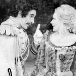 A black-and-white photo of a man and woman in historical costumes. The man, with a mustache and curly hair, points his finger while smiling at the woman, who has curly blonde hair and is also smiling. Both appear engaged in an animated conversation.