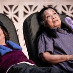 Two women lying on lounge chairs, the woman on the left is smiling and wearing a purple top and blue hat, the woman on the left is frowning and wearing a purple nightdress and holding a purple electric fan to her face.