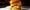 A juicy cheeseburger with melty cheese, lettuce, pickles, and mayonnaise is served on a shiny brioche bun on a parchment-lined tray. The background is softly blurred, emphasizing the well-lit, appetizing burger in the forefront.