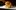 A juicy cheeseburger with melty cheese, lettuce, pickles, and mayonnaise is served on a shiny brioche bun on a parchment-lined tray. The background is softly blurred, emphasizing the well-lit, appetizing burger in the forefront.