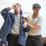 Two men are in a room. One is wearing a blue jacket, grey shirt, and drinking from a glass, looking unsteady. The other man, in a flat cap and striped shirt, is behind him, with one hand on the first man's arm and the other on his shoulder, appearing to support him.