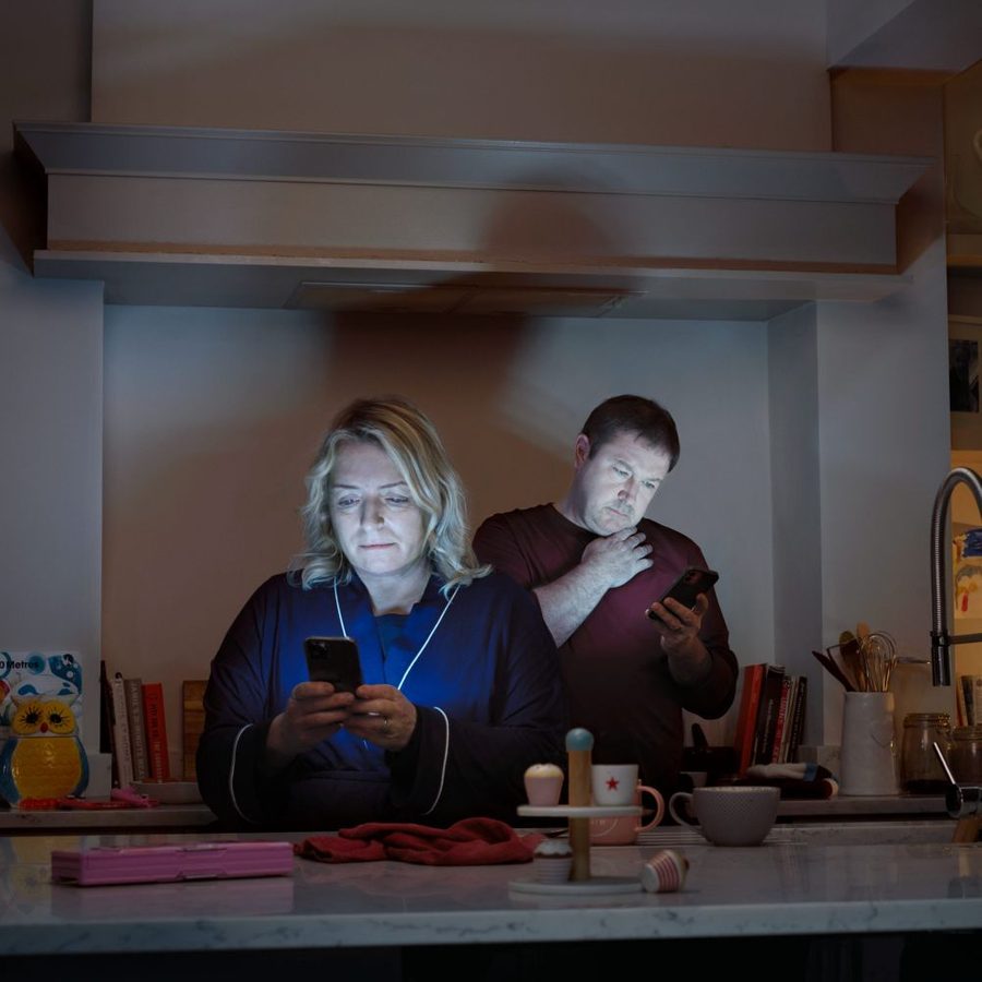 middle-2000x1000-masthead A woman and a man stand in their kitchen, each engrossed in the screens on their mobile phones.