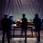 A naked man lies on a table under a dim spotlight, surrounded by five blurry, silhouette-like figures in motion. The background is dark and indistinct, with a bed on the left and a piece of equipment on the right.