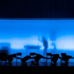 Six silhouetted figures bow low on a dark stage with chairs in front of a transparent blue-lit curtain. The shadow of a person holding a long walking stick is visible through the curtain.