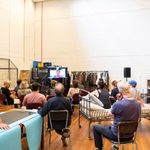 17 people in rehearsals, sat on chairs and a bed. They are all on a call with a man who is displayed on a large screen that has been put in front of them.