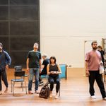 Six actors in a rehearsal room moving around the space to practice a scene. A woman in the centre is sat down and holding a white teacup and saucer.