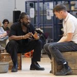 Two men are engaged in an intense conversation. They are seated on wooden crates, with one gesturing expressively. In the background, other people are seated, observing.