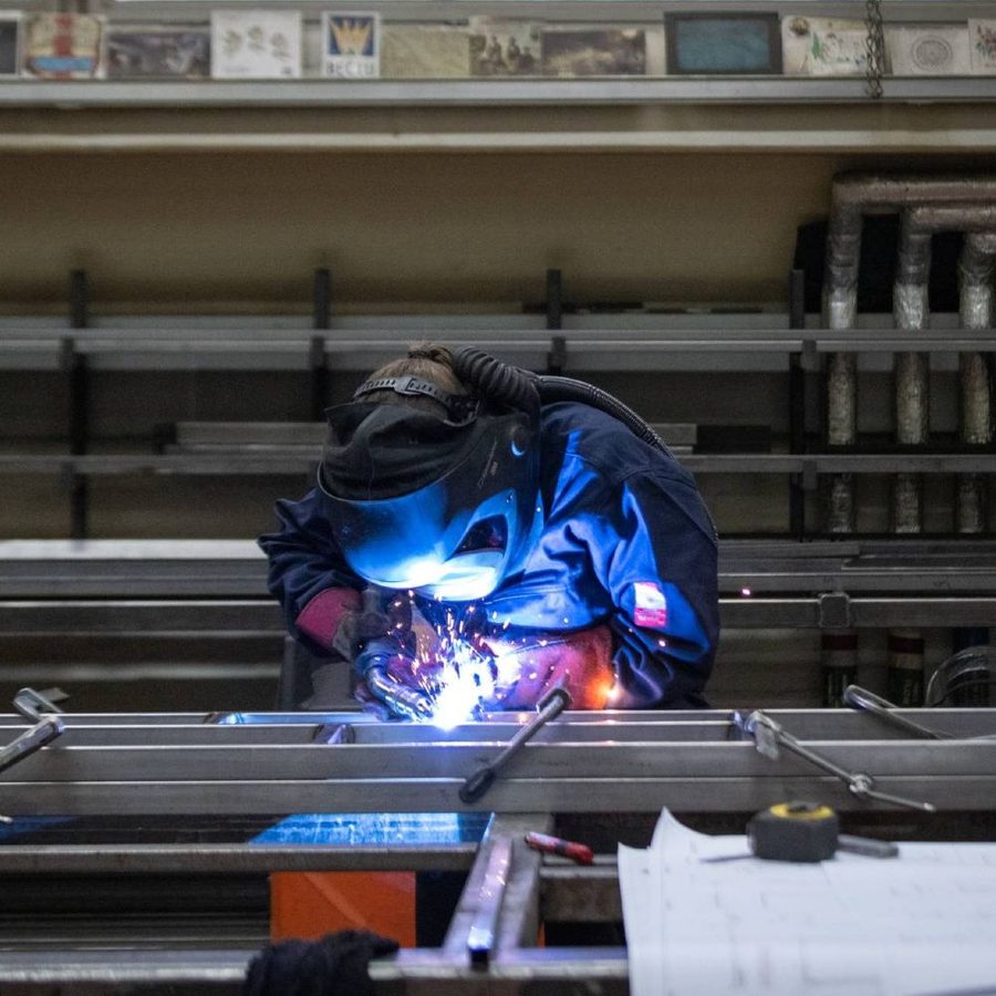 National-Theatre-Apprentice-welding-2023-masthead-CameronSlaterPhotography A person wearing protective gear and a welding helmet is performing welding work on a metal structure in a workshop. Various tools, pipes, and sheets are visible around the workspace.