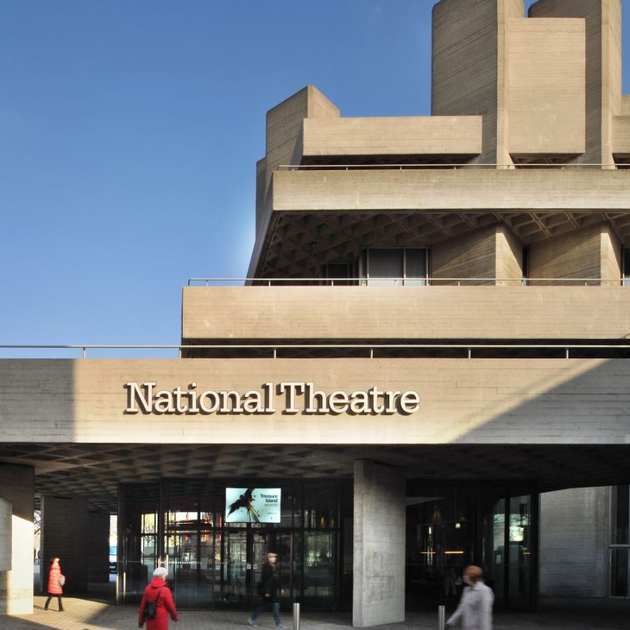 National-Theatre-entrance-pavillion-foyer-2000x1000-photo-phillip-vile A modern, spacious lobby with a revolving glass door, soft seating, and people walking through. Concrete walls and ceiling feature geometric patterns, and there's a digital directory on the right wall. Bookstore shelves are visible in the background.