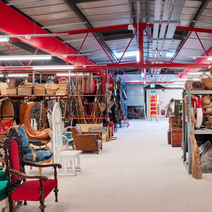 national-theatre-green-store-2000x1250-photo-pete-goding-006-aspect-ratio-2000-1000 A warehouse filled with assorted vintage furniture, baskets, and various items. Red overhead pipes are visible along with a ladder in the background. Shelves are stocked with a variety of eclectic objects including chairs, statues, and decorative items.
