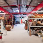 A warehouse filled with assorted vintage furniture, baskets, and various items. Red overhead pipes are visible along with a ladder in the background. Shelves are stocked with a variety of eclectic objects including chairs, statues, and decorative items.