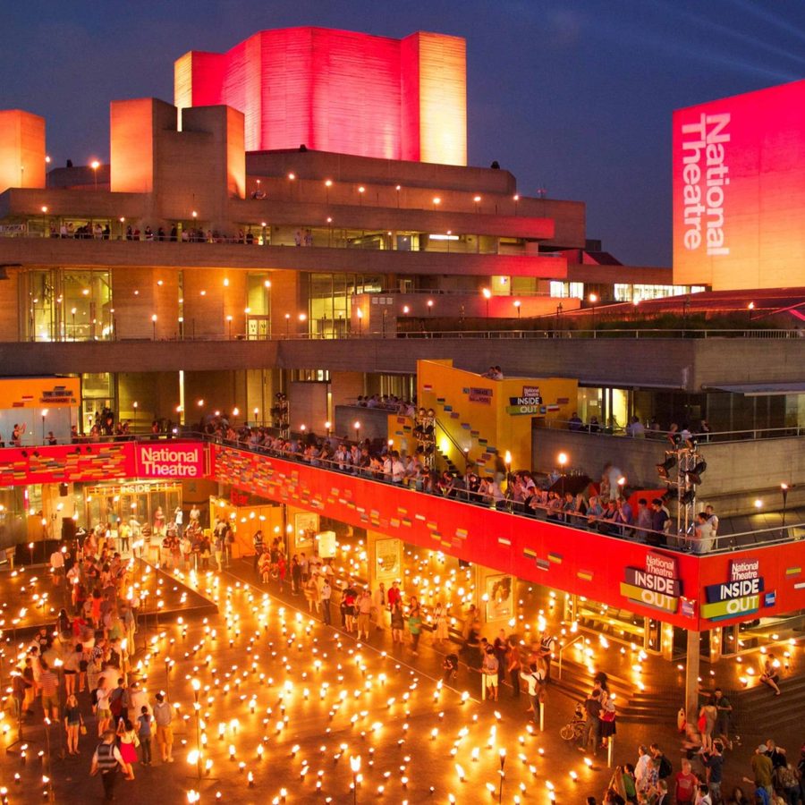 A brightly lit National Theatre at night, featuring warm orange and red hues. The exterior is decorated with numerous lights, and people are gathered outside. The building's modern architecture is highlighted under the evening sky.