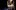 Image provided by Creative & Cultural Skills© Briony Campbell” A person stands on scaffolding, repairing or adjusting a hanging light fixture. Below, another person watches from the ground. The setting is a stage with a rustic wooden structure under theatrical lighting.
