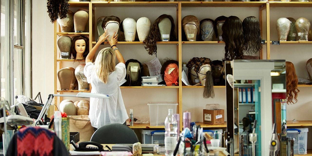 A person arranges wigs on a shelf in a room filled with various wig mannequins, styling heads, and hairpieces. The workspace is filled with styling tools, bottles, and hair products, creating a busy, organized environment.