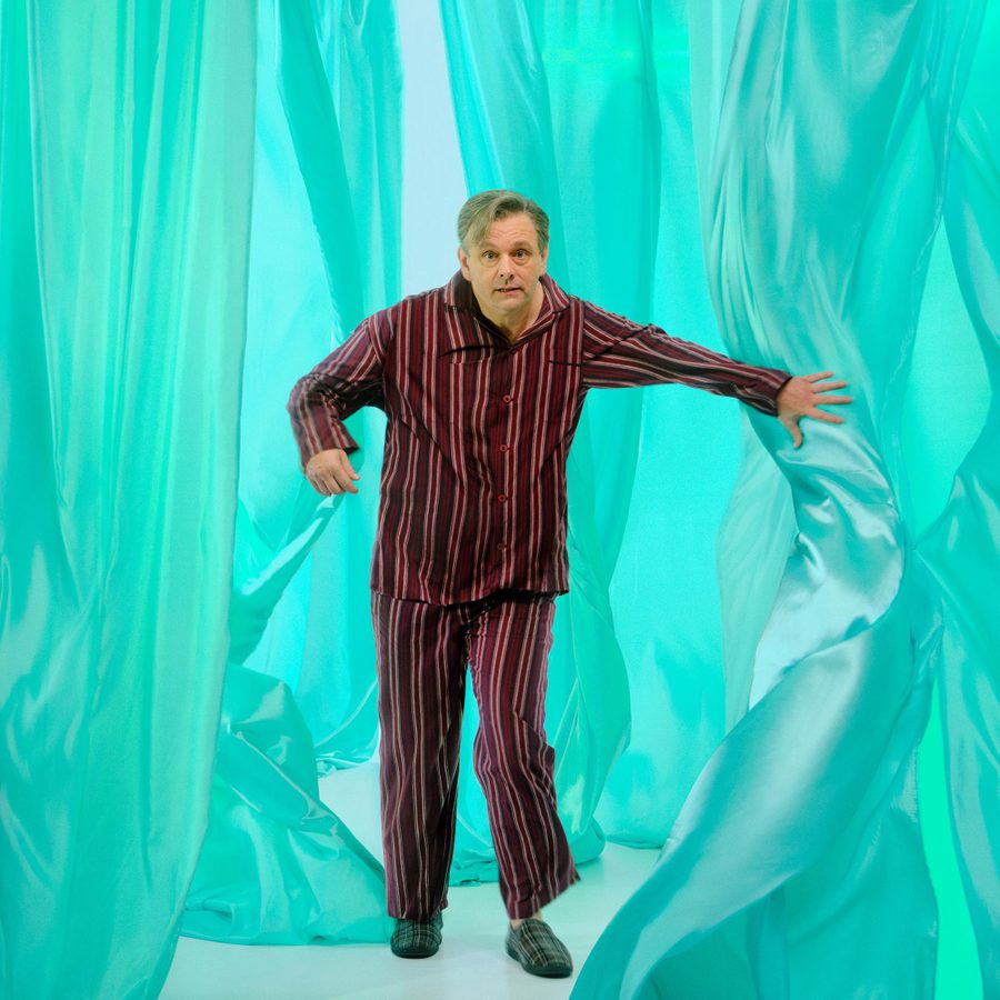 Nye-Michael-Sheen-National-Theatre-2025-masthead A man wearing red and black striped pajamas and plaid slippers is moving through flowing, translucent teal curtains. He extends his left arm forward, to part one of the curtains. The background consists of soft, undulating teal curtain layers, creating an abstract, dreamy environment.