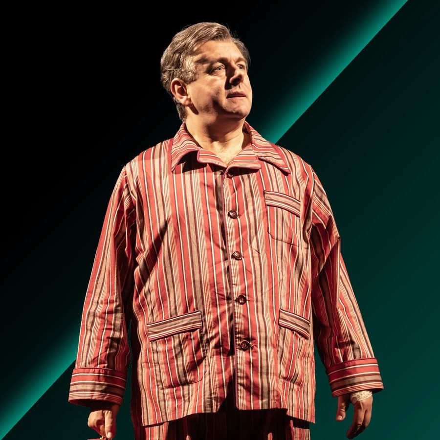Nye-Take-Your-Seats-National-Theatre-2000x1000 A person (Michael Sheen) wearing a striped pajama top stands against a bold, dark green and black gradient background, looking upward with a thoughtful expression.