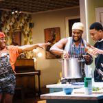 Three men are in a lively kitchen. One man in an orange shirt and camouflage overalls gestures energetically. Another in a yellow bandana and striped apron stirs a large pot. The third man, in a black T-shirt, looks attentively into the pot.