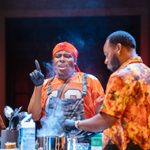 Two men are cooking in a kitchen. One man, wearing an orange shirt and black rubber gloves, is gesturing and speaking passionately. The other man, in an orange patterned shirt, is focused on stirring a pot that is emitting steam. Various kitchen utensils and ingredients are on the counter.