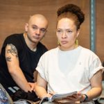 A woman with a focused expression is holding a notebook and sits in the foreground, while a man with a shaved head and tattooed arm looks on from the background in a room with wooden panels.