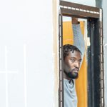 A man with a solemn expression peeking through a partially built doorframe surrounded by light grey walls, in a set under construction.
