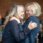 Two women joyfully embrace, smiling closely at each other, in a warmly lit indoor setting, with a blurry background of people and chairs.