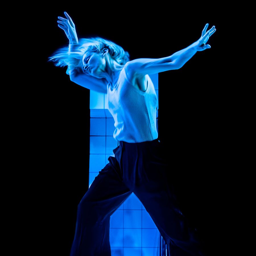 People-Places-and-Things-v2-2000x1000px A woman dances, wearing a white sleeveless top and dark trousers.