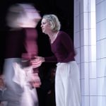A dynamic theater scene featuring two actors, one a blurred figure in motion, in expressive poses on a minimalist set, with an audience visible in the background.
